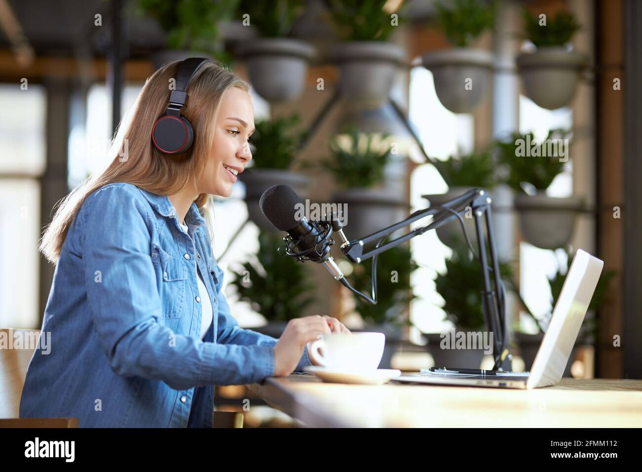 Vista laterale di una giovane donna sorridente e bella che lavora in un computer portatile con cuffie in un bar con un caffè gustoso. Concetto di comunicazione di processo in trasmissione diretta con le persone. Foto Stock