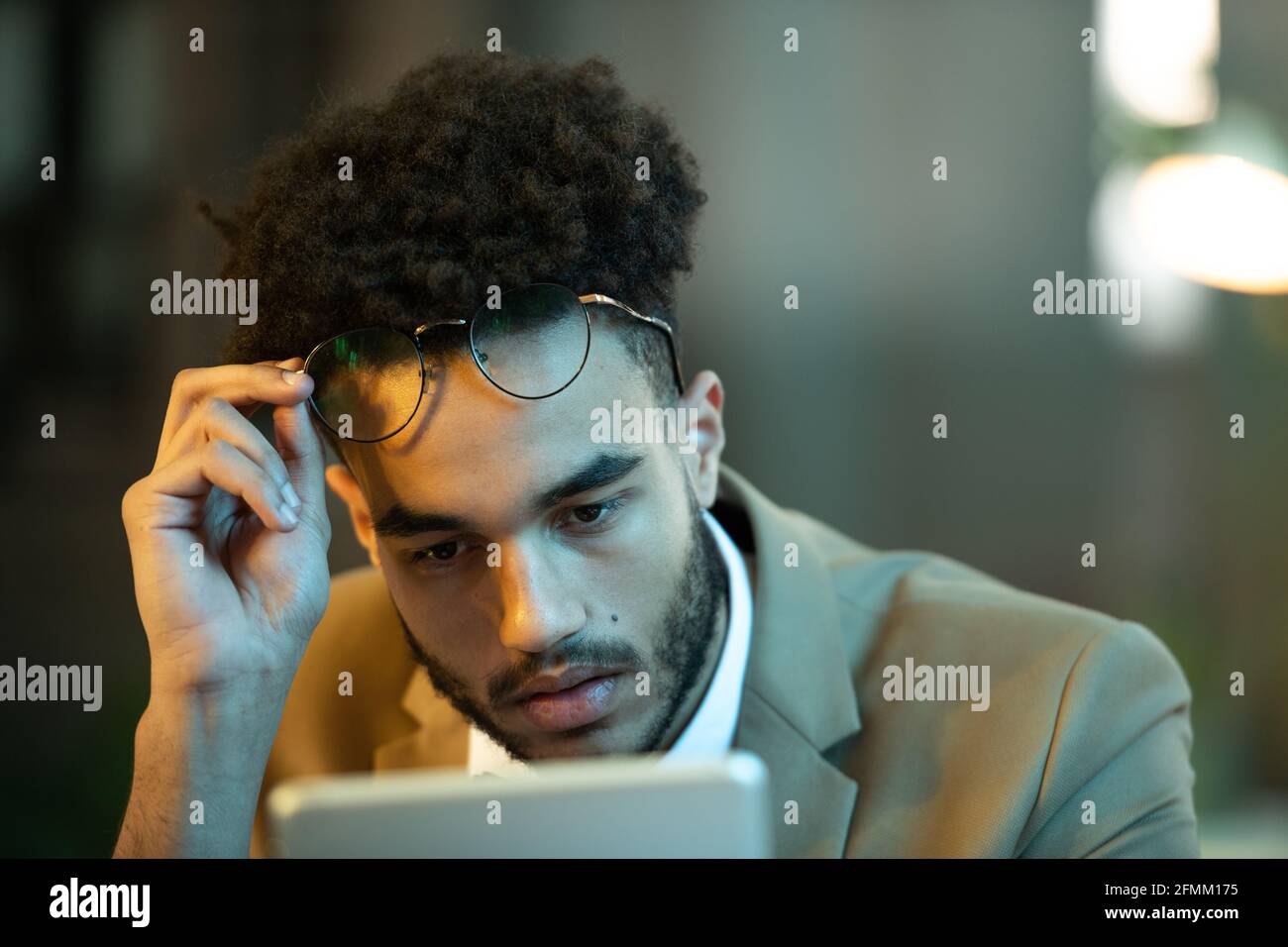 Occupato giovane manager nero con acconciatura afro che tira fuori gli occhiali mentre legge articolo su tablet, lavora tardi Foto Stock