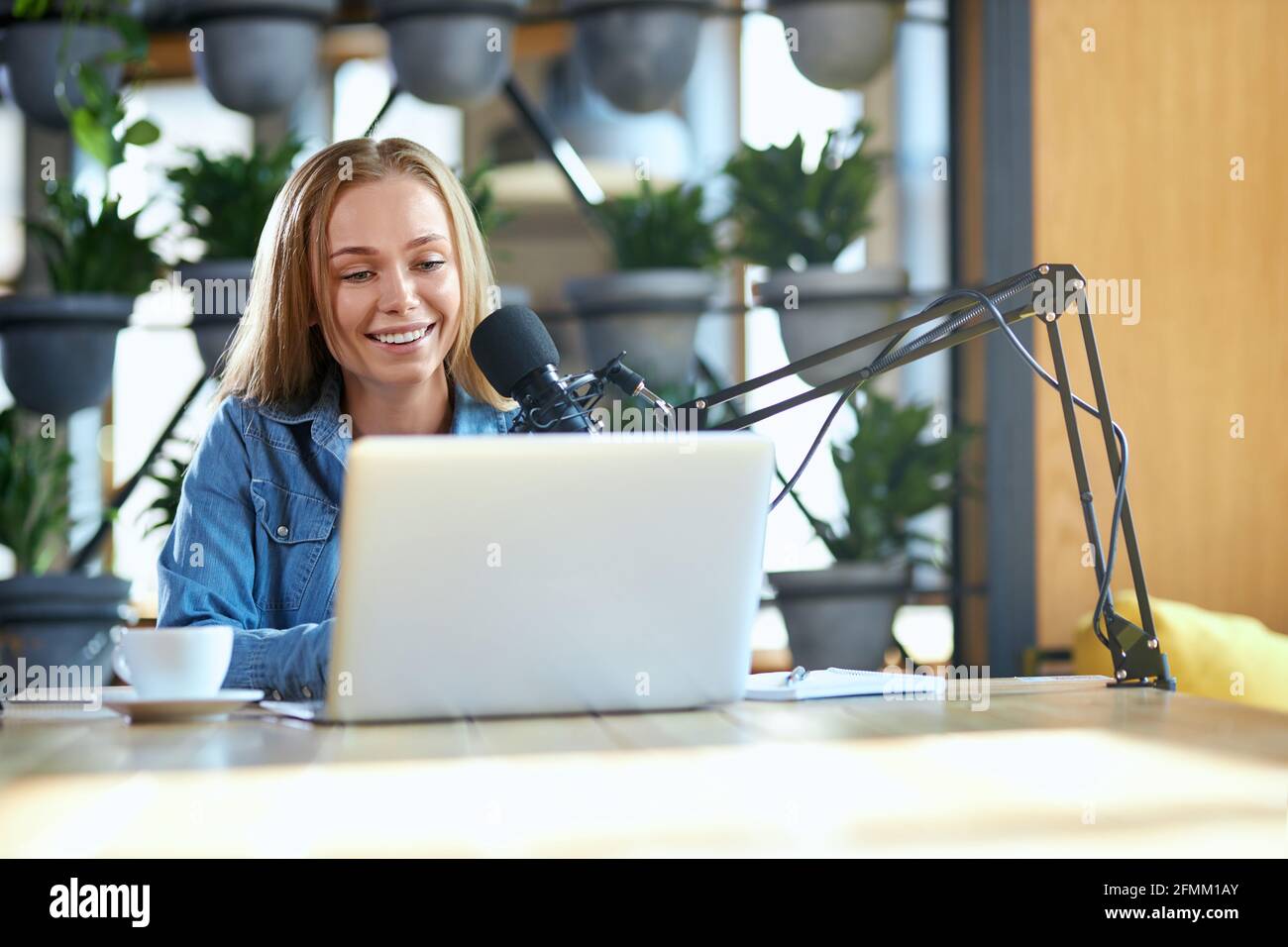 Vista frontale di una giovane donna carina con un buon umore che comunica online in un computer portatile con un microfono moderno nel bar. Concetto di processo che lavora su radio con microfono e computer portatile. Foto Stock