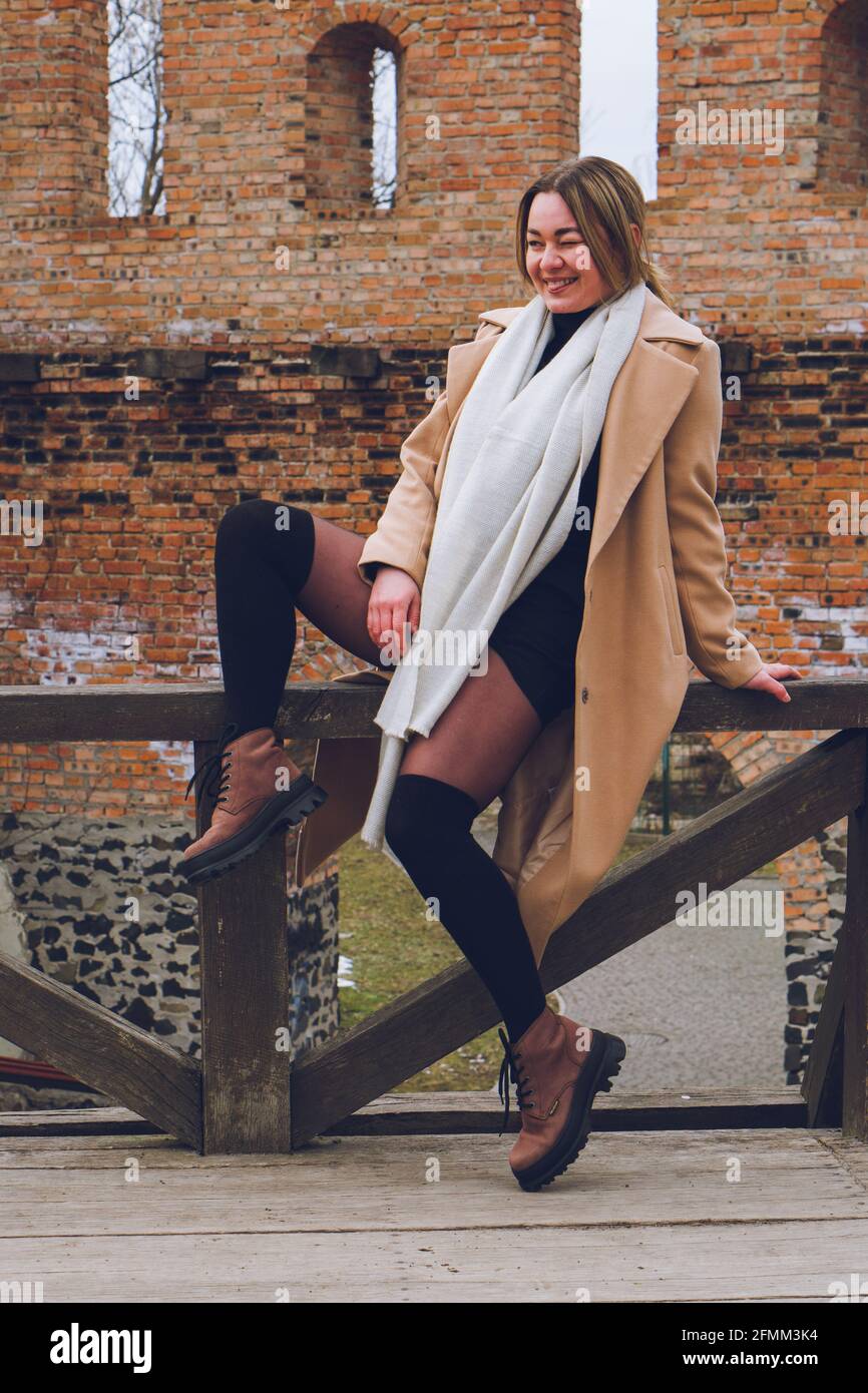 Bella giovane donna sorridente felice si siede su recinzione in un cappotto beige, sciarpa di cashmere di lana bianca e stivali marroni, fotografia di stile di strada. Rumore di pellicola, così Foto Stock