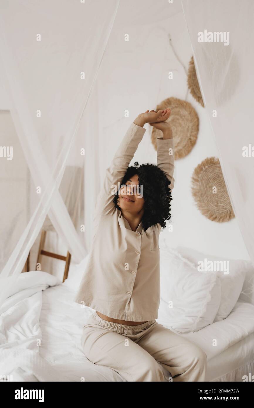 Donna afroamericana in sleepwear che si allunga e sorride con occhi chiusi seduti a letto a casa. Foto Stock