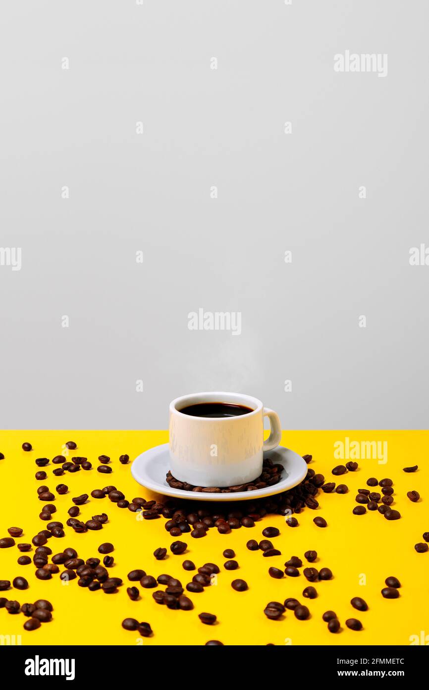 Tazza di caffè sopra il tavolo giallo luminoso e sfondo grigio e molti chicchi si sono rovesciati intorno ad esso. Spazio di copia. Composizione verticale. Foto Stock