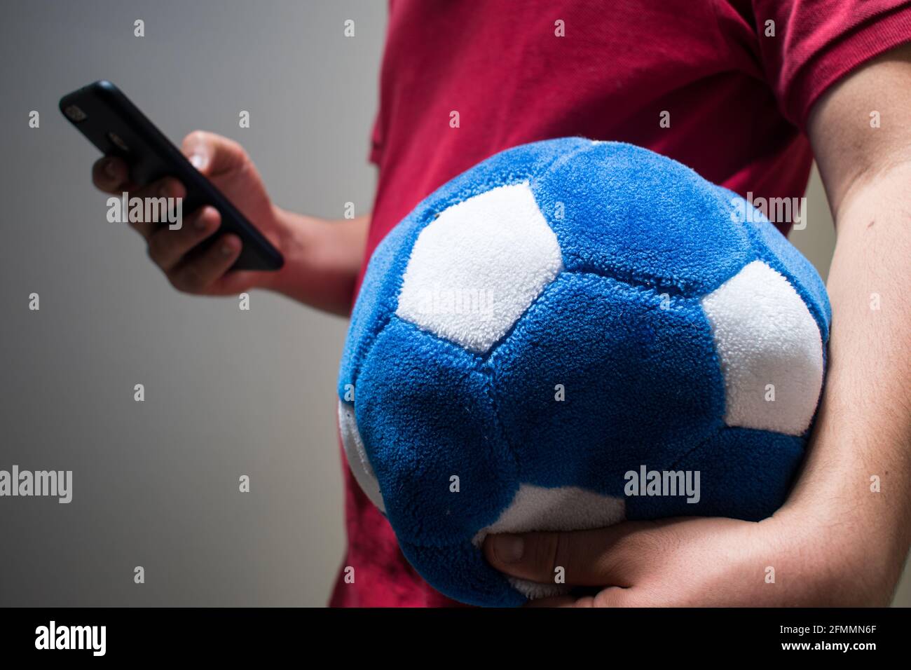 Uomo che indossa una t-shirt rossa guardando il suo cellulare e tenendo una palla di calcio blu Foto Stock