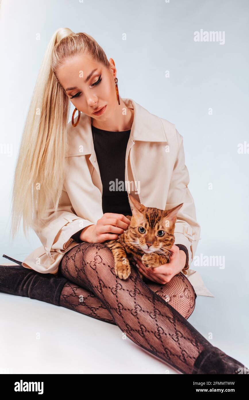 Ritratto di una giovane bella donna bionda con un gatto bengala. Una bella ragazza seduta sul pavimento guarda il gatto, elegante acconciatura, cheeky Foto Stock