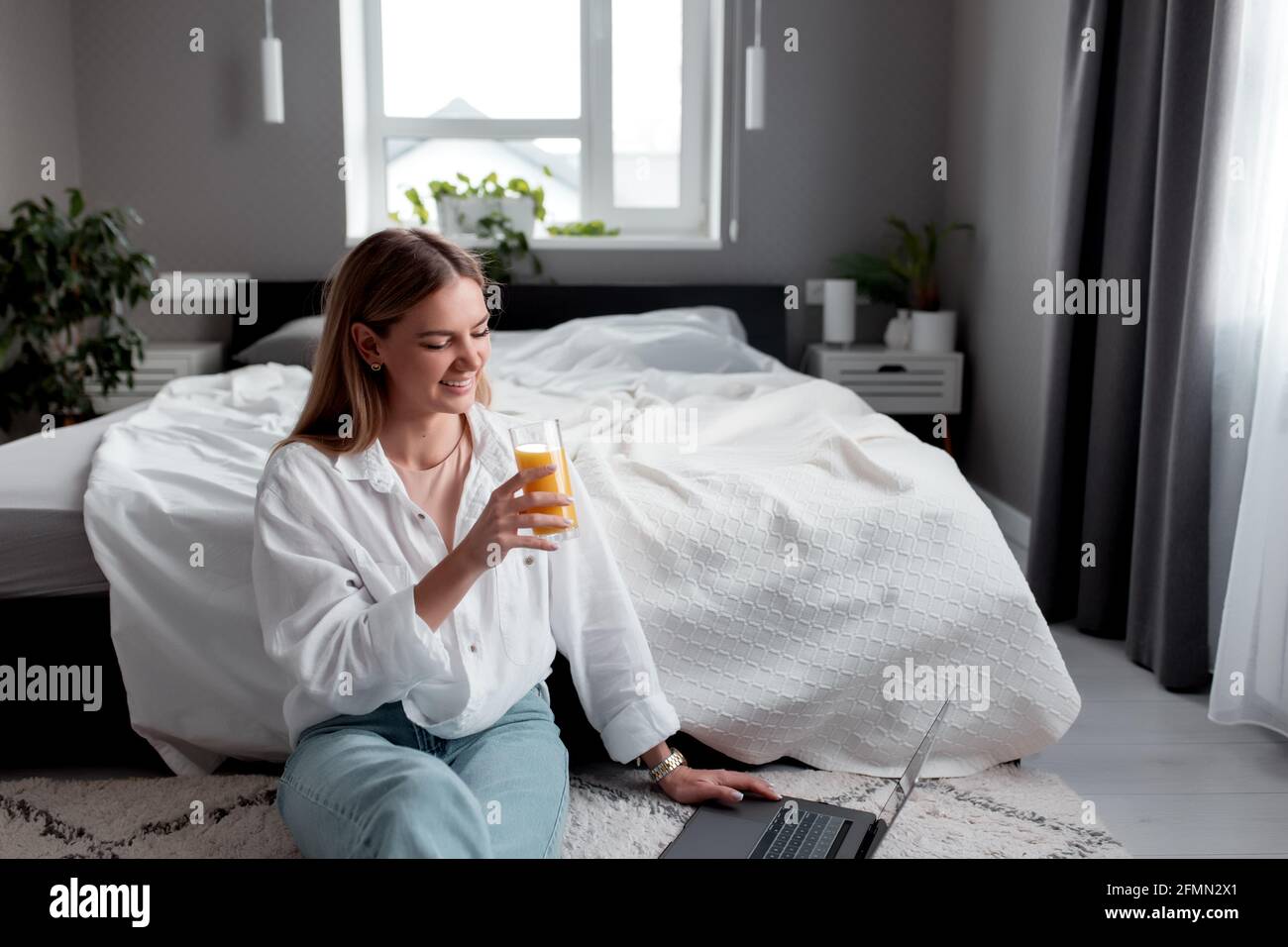 Bella ragazza caucasica sorridente si siede sul pavimento in camera da letto e utilizzare il computer portatile per il lavoro remoto e bere succo d'arancia. Comunicazione, tecnologia, telecomando WO Foto Stock