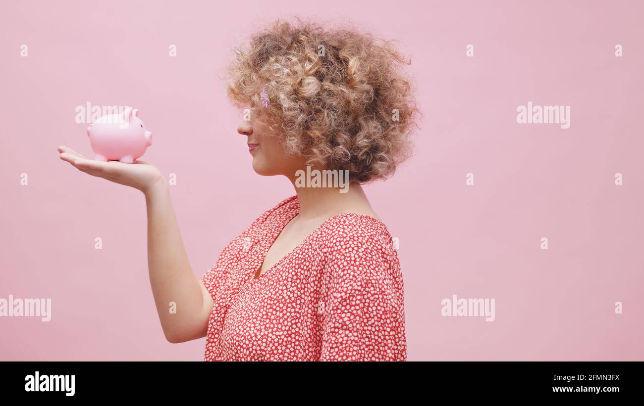 Donna che tiene una piccola banca piggy in mano e che fa facce divertenti. Grande aspettativa di crescita degli investimenti di piccole dimensioni. Foto di alta qualità Foto Stock