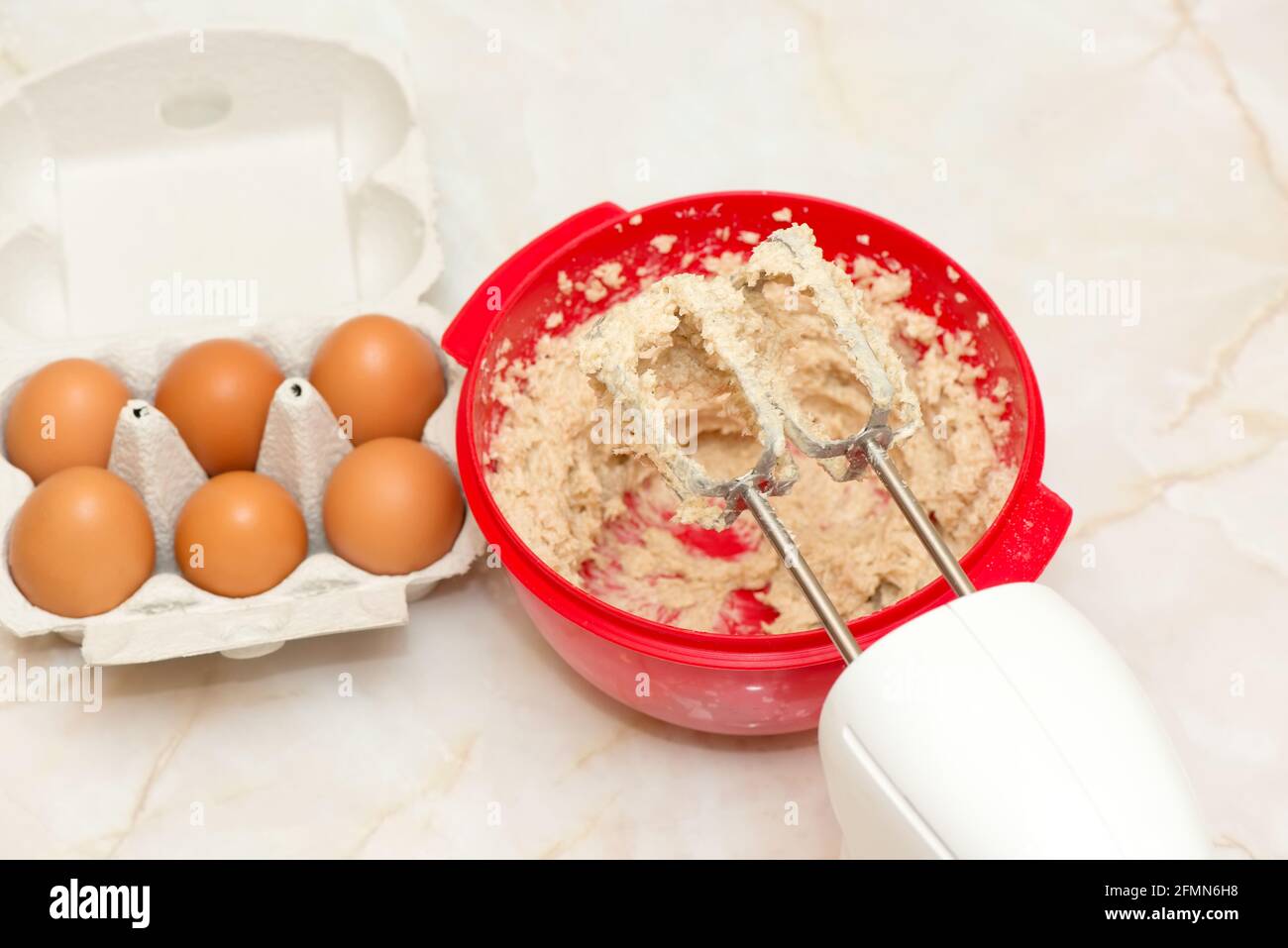 Frusta elettrica su una ciotola rossa con impasto e uova cartone Foto Stock