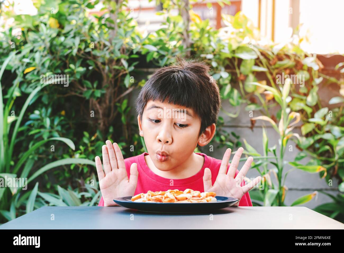 Carino bambino asiatico in una camicia rossa ha mostrato una buona espressione quando ha visto una pizza in un piatto posto di fronte al tavolo. Foto Stock