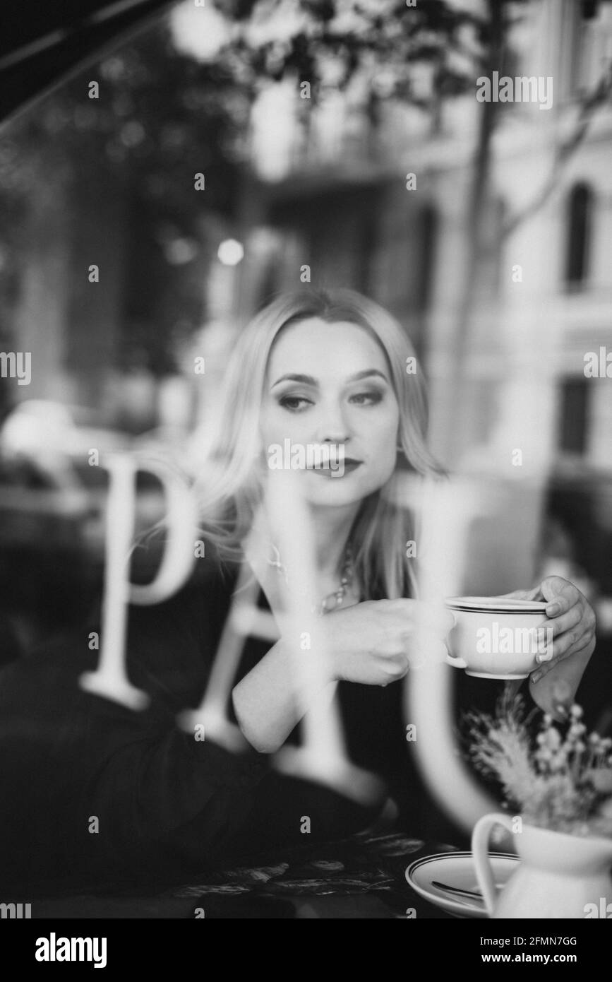 Donna bionda felice in un caffè con una tazza di caffè aromatico, foto attraverso la finestra di vetro. Messa a fuoco selettiva morbida, rumore artistico. Foto Stock