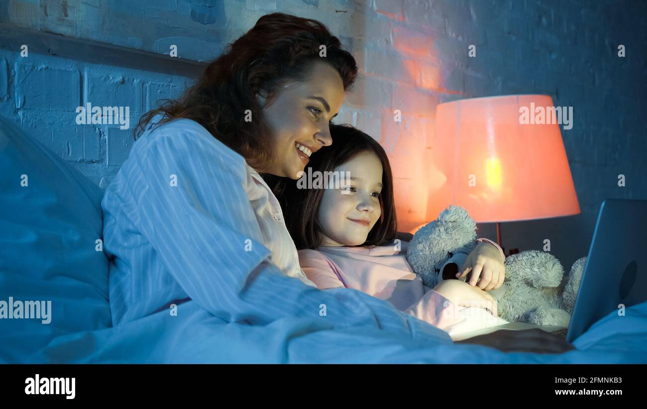 Donna sorridente che usa un computer portatile vicino alla figlia con un giocattolo morbido letto Foto Stock Donna sorridente che usa un computer portatile vicino alla figlia con un giocattolo morbido letto Foto Stock