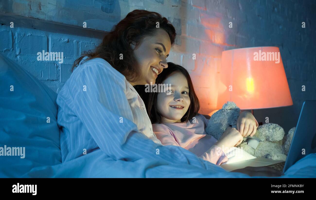 Donna in pajama usando un computer portatile vicino sorridendo figlia con morbido giocattolo sul letto Foto Stock Donna in pajama usando un computer portatile vicino sorridendo figlia con morbido giocattolo sul letto Foto Stock