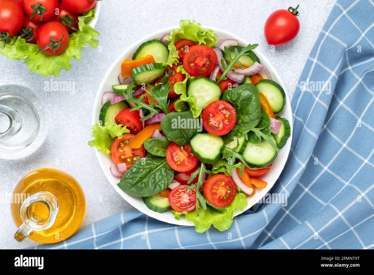 Insalata di verdure estive con pomodori ciliegini, cetrioli, rucola, basilico e condita con olio d'oliva. Tovagliolo a scacchi blu su un tavolo grigio. Vista da a. Foto Stock