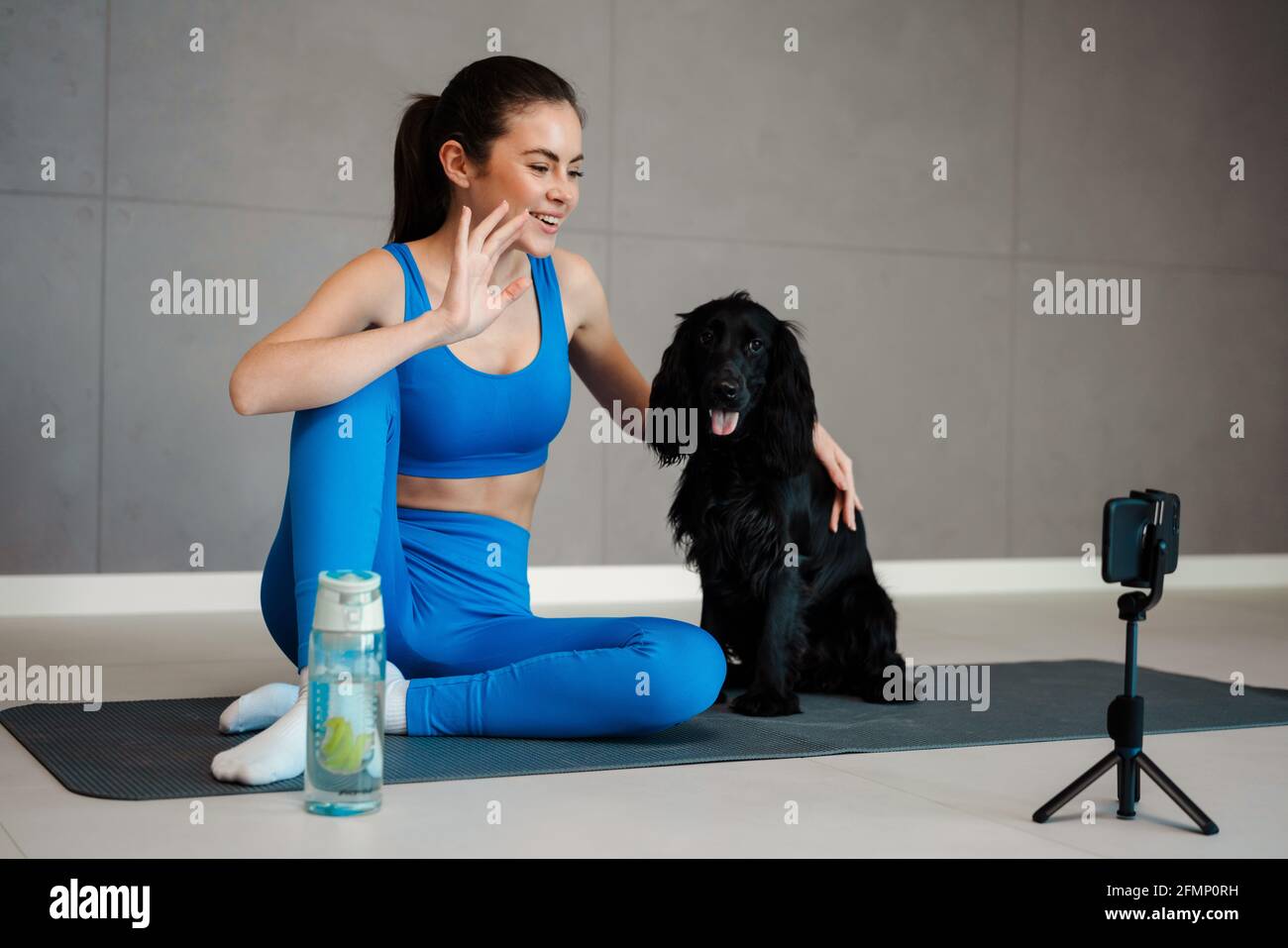 Felice giovane donna bruna blogger esercizio con il suo cane su un tappetino fitness davanti alla fotocamera all'interno Foto Stock