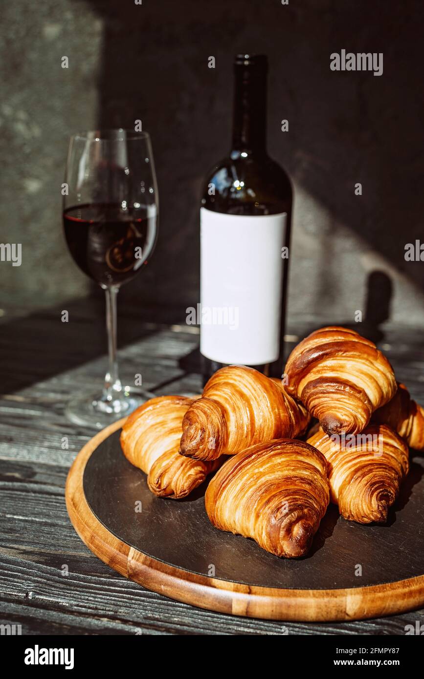 Croissant profumati appena sfornati su un pannello di pietra nera, un bicchiere di vino rosso e una bottiglia. Cena romantica. Foto Stock