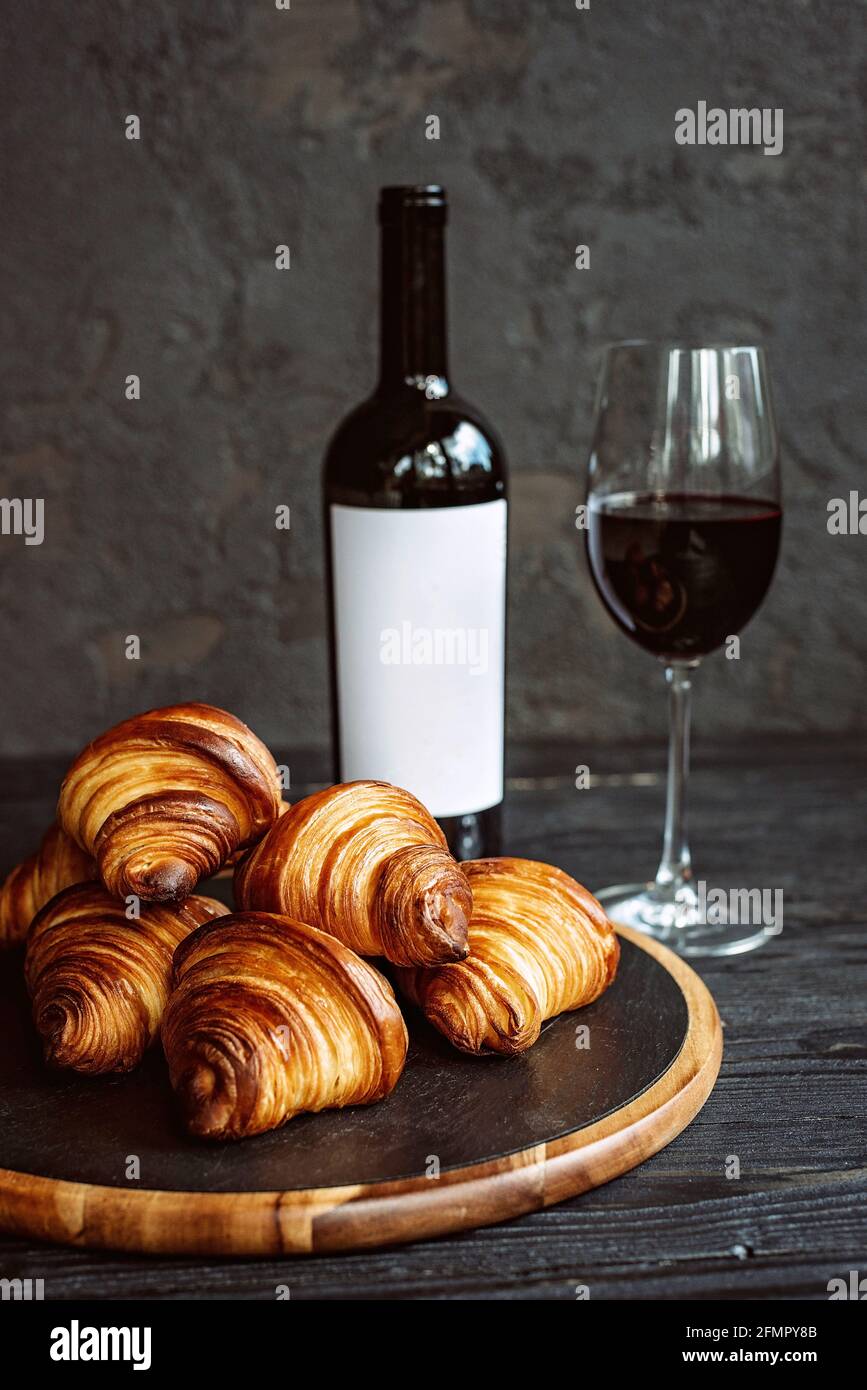 Croissant profumati appena sfornati su un pannello di pietra scura, un bicchiere di vino rosso e una bottiglia. Cena romantica. Foto Stock