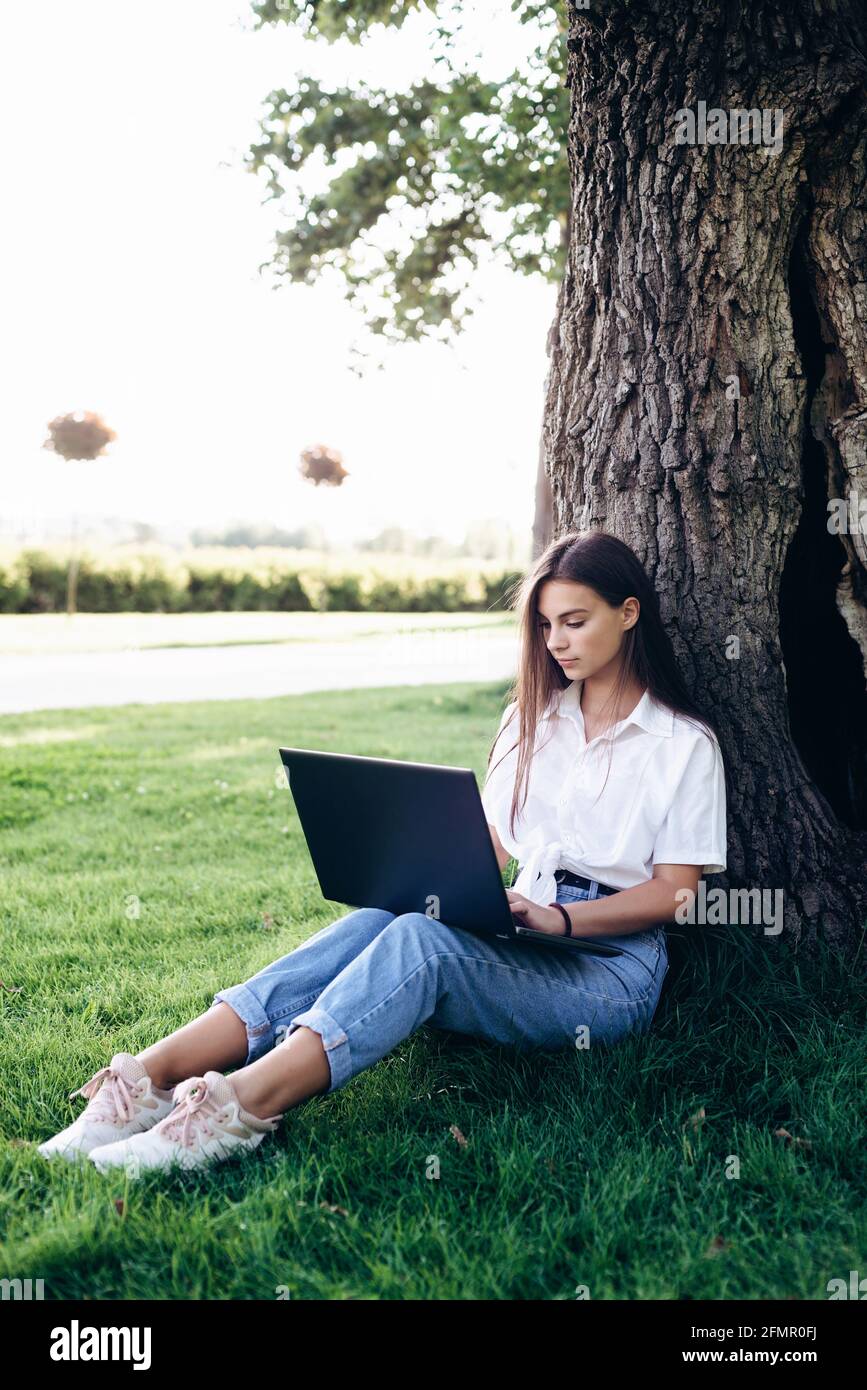 la studentessa con un computer portatile all'aperto si siede sull'erba del parco, navigando in internet o preparandosi agli esami. Tecnologia, istruzione e lavoro a distanza Foto Stock