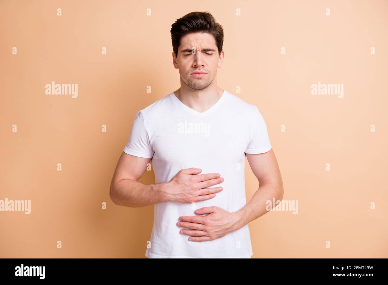 Foto del giovane uomo occhi chiusi che tengono le mani sul ventre si sente male isolato su sfondo di colore beige Foto Stock