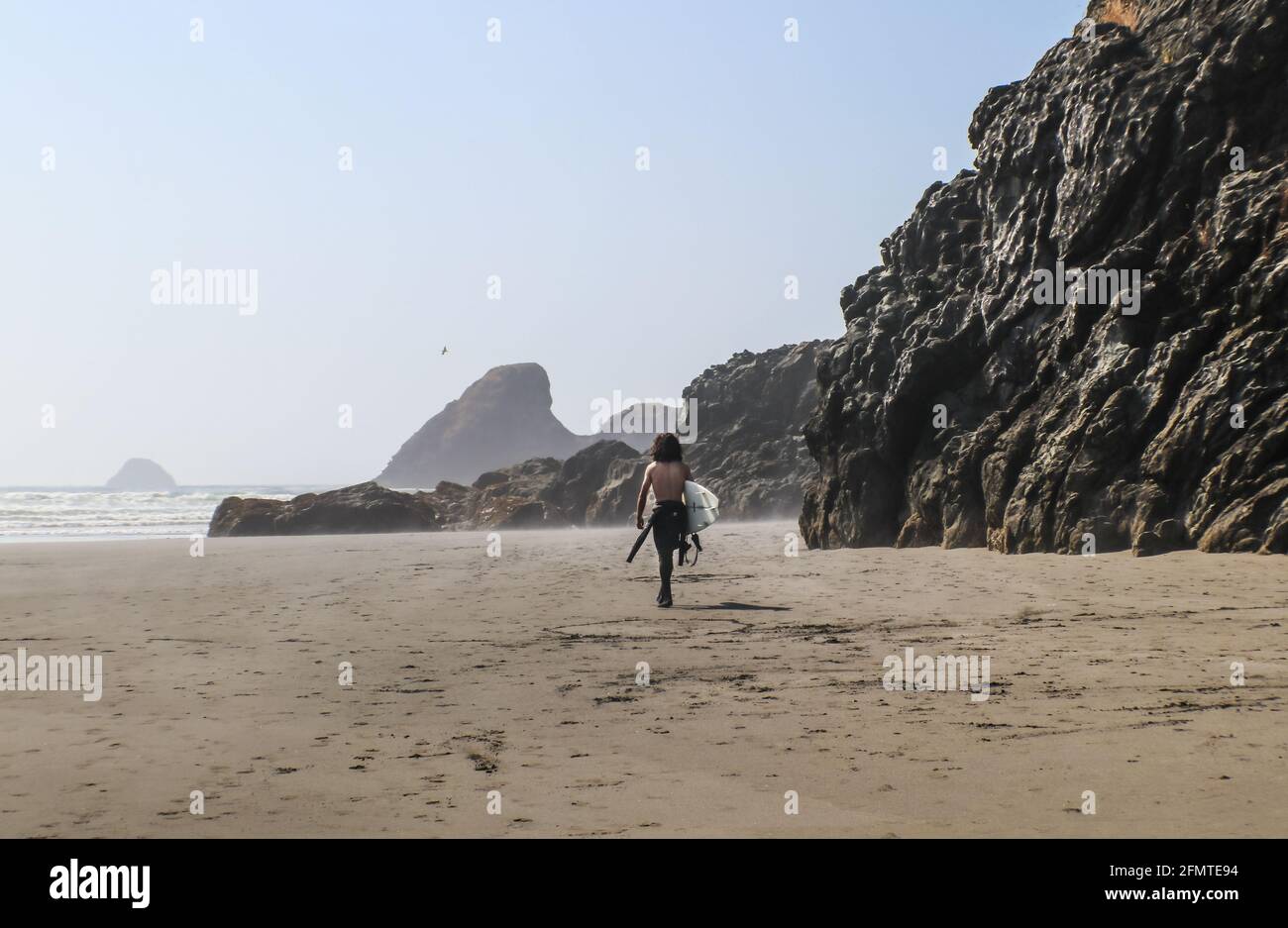 Giovane uomo surfer senza camicia e bufy lungo scuro i capelli camminano lungo la spiaggia nebbiosa con torreggianti scogliere di roccia bagnata verso l'oceano con le onde che rotola dentro - shad Foto Stock