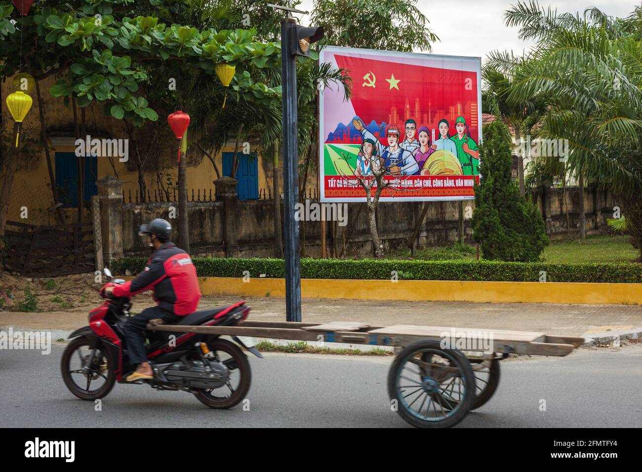 Moto vietnamita passando manifesto di propaganda comunista sul lato della strada, Hoi An, Vietnam Foto Stock