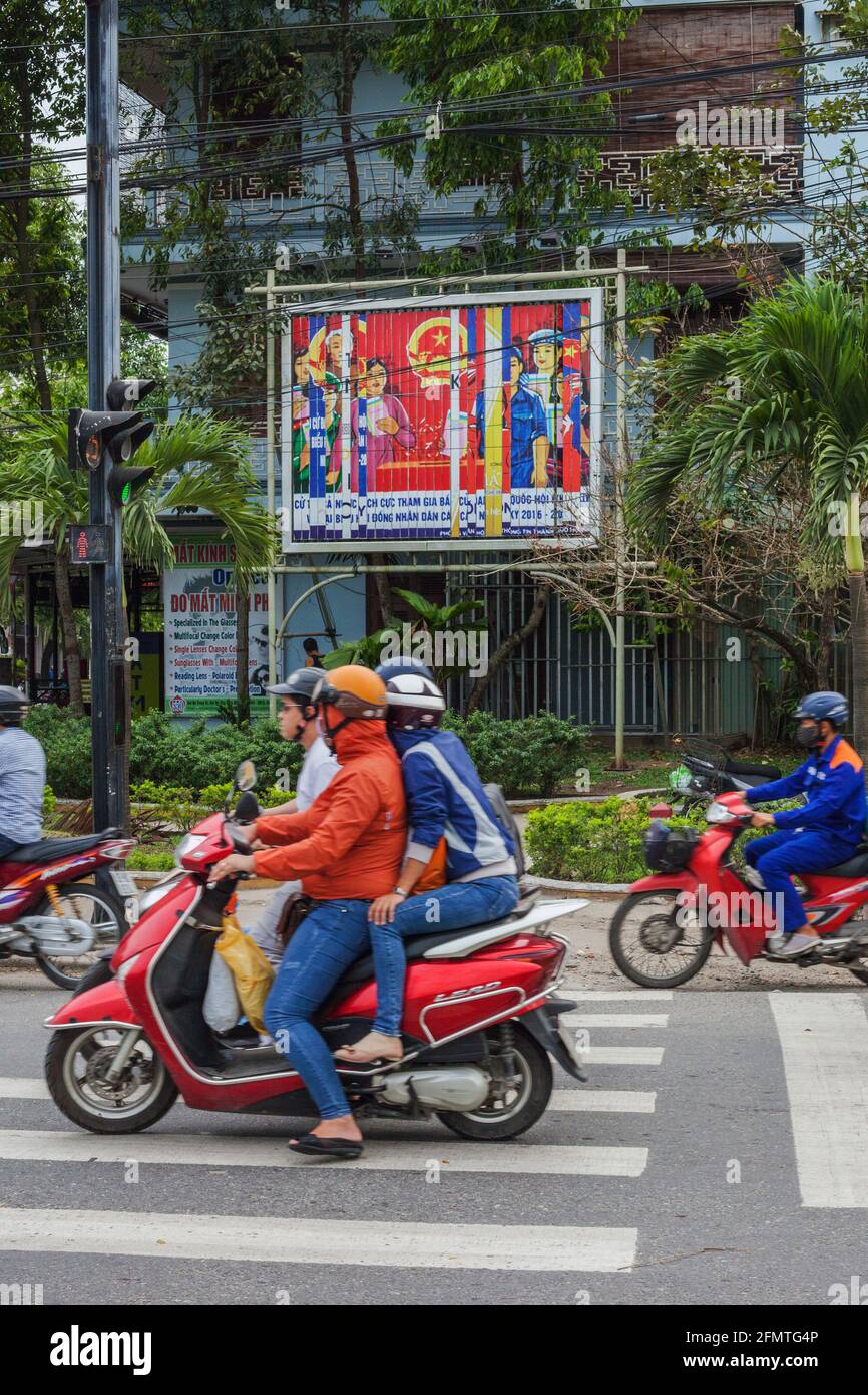 Gli automobilisti vietnamiti che passano manifesto di propaganda comunista sul lato della strada, Hoi An, Vietnam Foto Stock