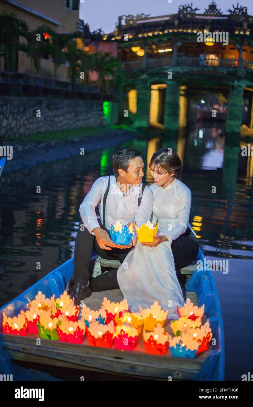 La coppia vietnamita sedette in barca di fronte al ponte giapponese rilasciare lanterne galleggianti colorati nel fiume, Hoi An, Vietnam Foto Stock