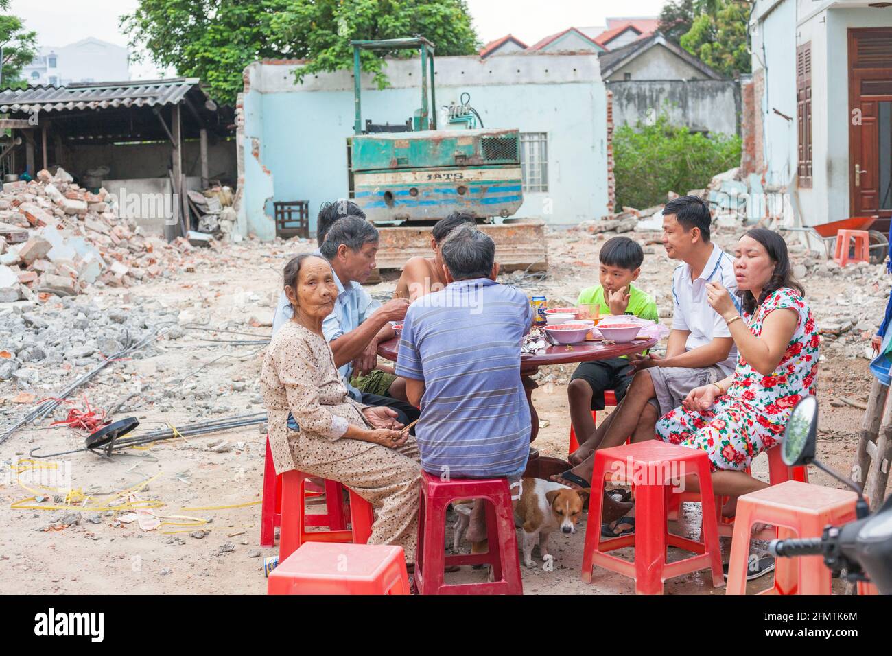 Famiglia vietnamita che ha pasto al tavolo fuori sul terreno di rifiuti con digger in background, Hoi An, Vietnam Foto Stock