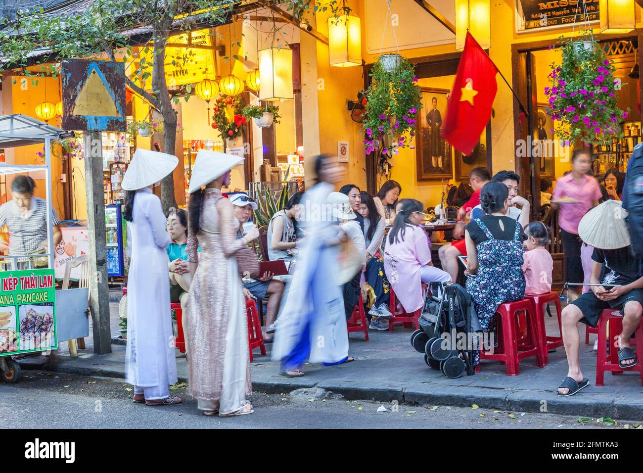 Graziose femmine vietnamite che indossano cappelli conici e abiti tradizionali di ao dai si trovano all'esterno del ristorante nella città vecchia, Hoi An, Vietnam Foto Stock