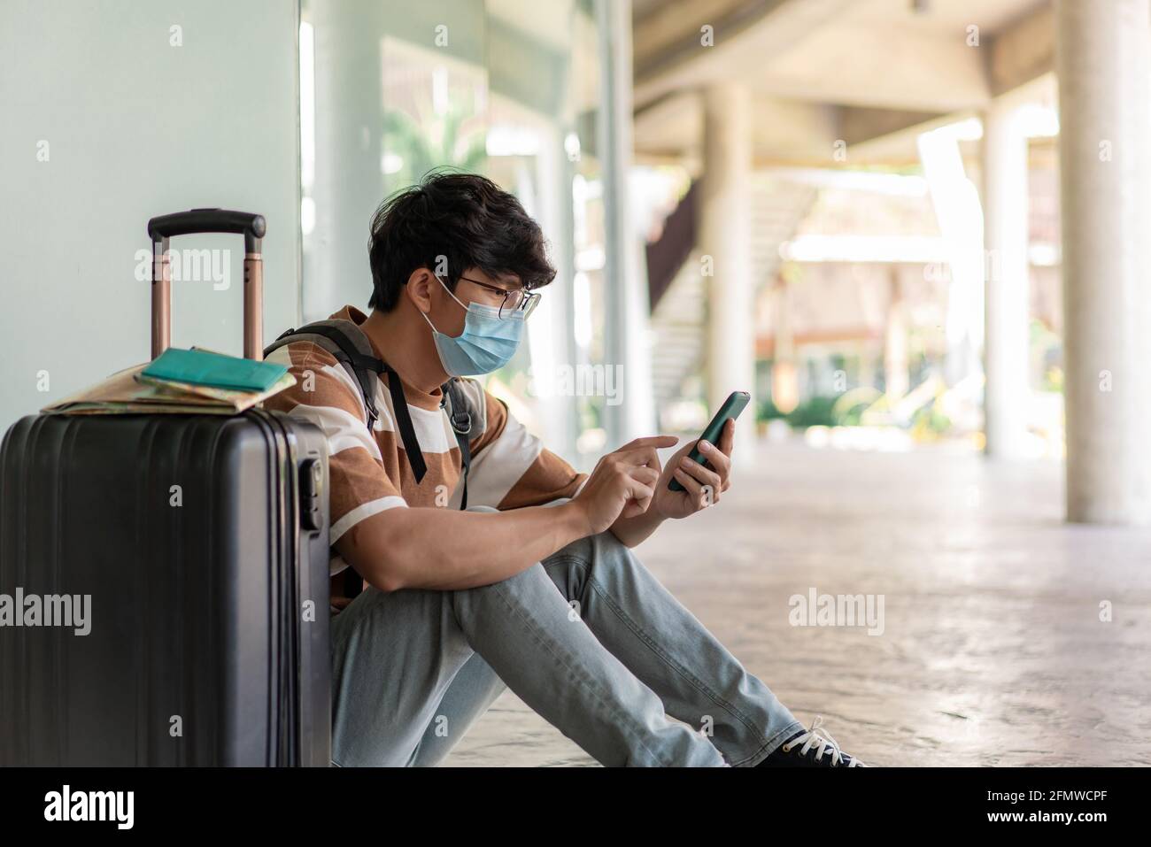 Viaggi. Un uomo che indossa occhiali con una camicia a righe e uno zaino, indossa una maschera, siediti sul pavimento. Utilizzando un telefono durante il viaggio con mappa, notebook sulla su Foto Stock