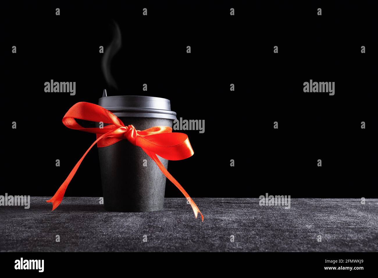 Tazza monouso per bevande da asporto in carta nera con vapore sul coperchio con prua in nastro rosso regalo al buio superficie e sfondo nero Foto Stock