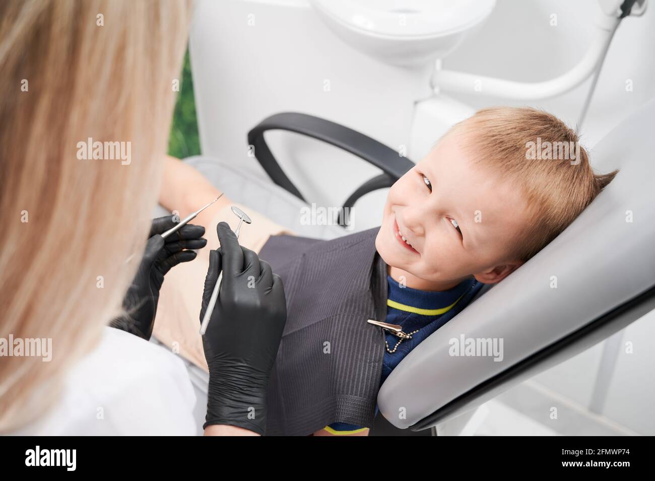 Adorabile ragazzo maschile guardando la macchina fotografica e sorridendo mentre dentista femminile tenendo specchio dentale ed esploratore in clinica dentale. Concetto di odontoiatria pediatrica e cura dentale. Foto Stock