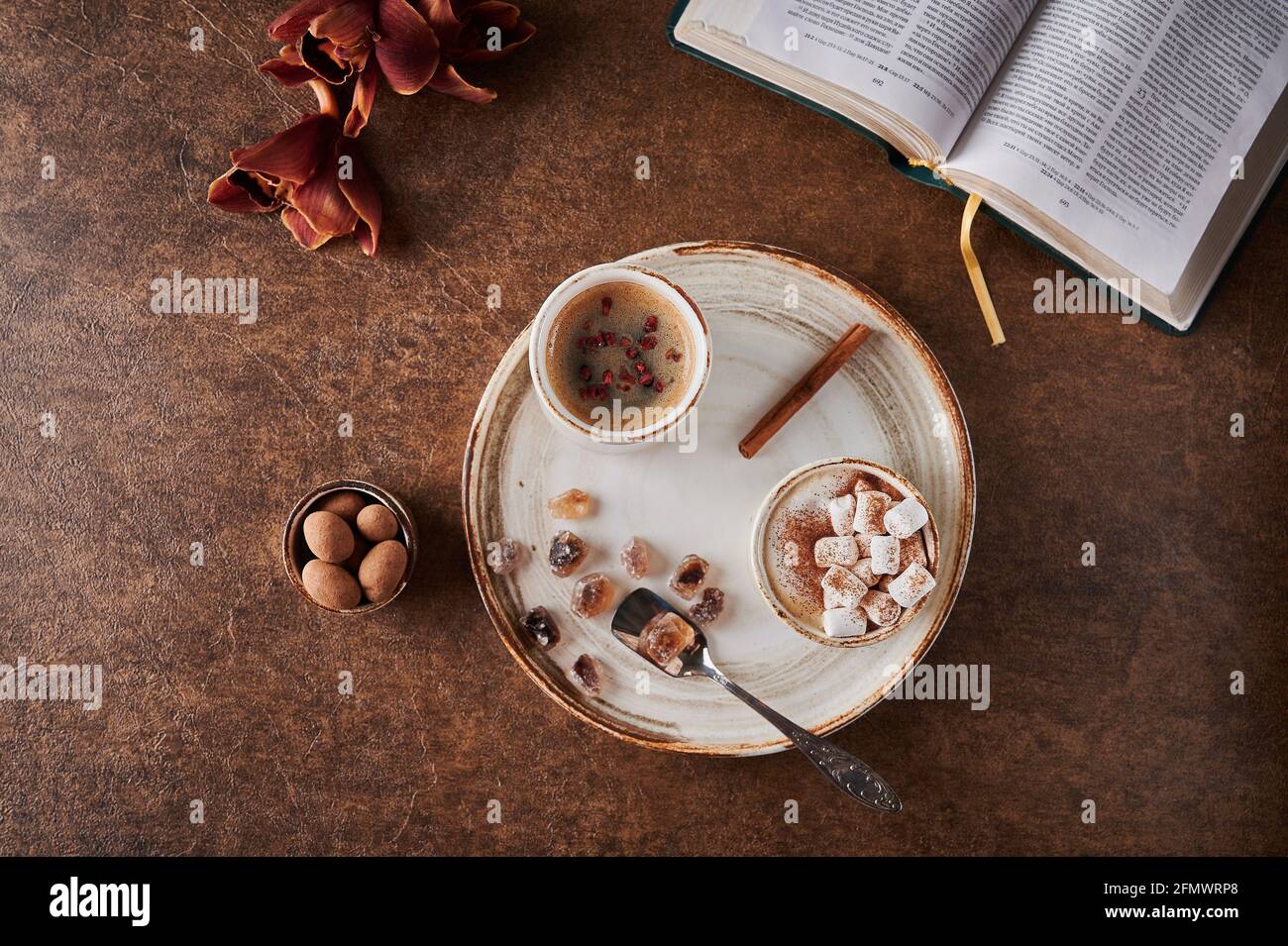 Vista dall'alto due tazze di caffè con schiuma e marshmallow e lamponi sublimati con cannella su piatto leggero con zucchero caramellato e cucchiaio. Nelle vicinanze si trova un libro aperto, fiori e caramelle Foto Stock