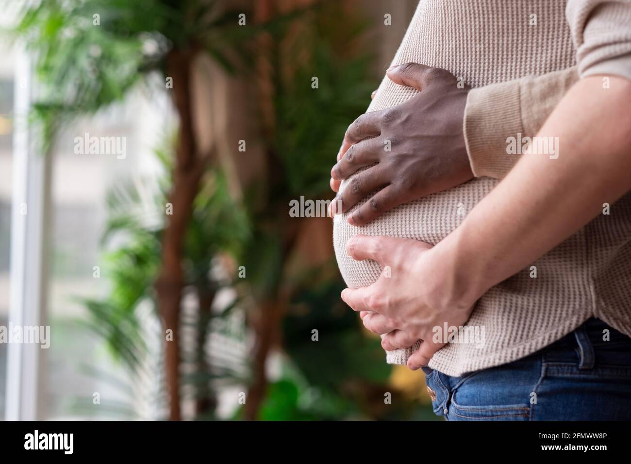 Il padre nero in attesa che tiene le mani sul ventre della sua moglie caucasica incinta. Matrimonio misto. Foto Stock