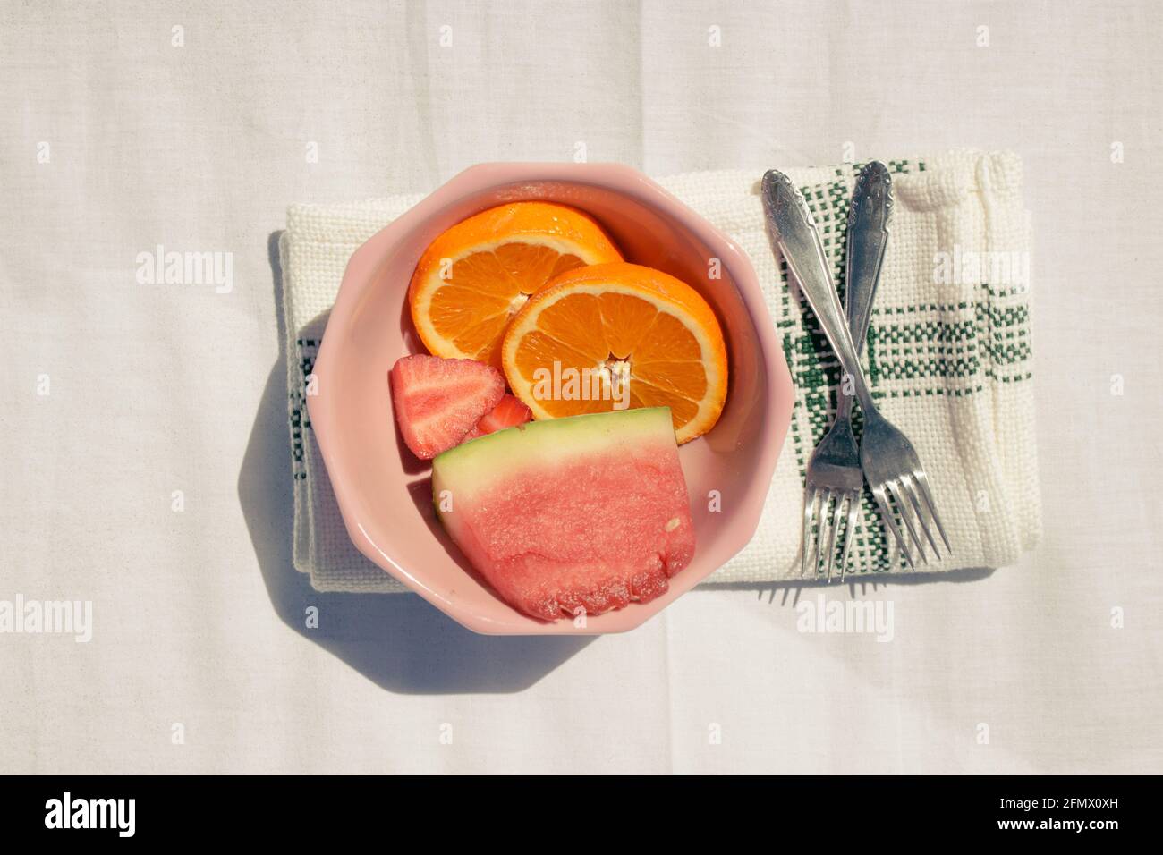 Immagine dettagliata in orizzontale di frutta assortita (arance, anguria e fragola) in una ciotola rosa insieme a due forchette su asciugamano in tessuto bianco Foto Stock
