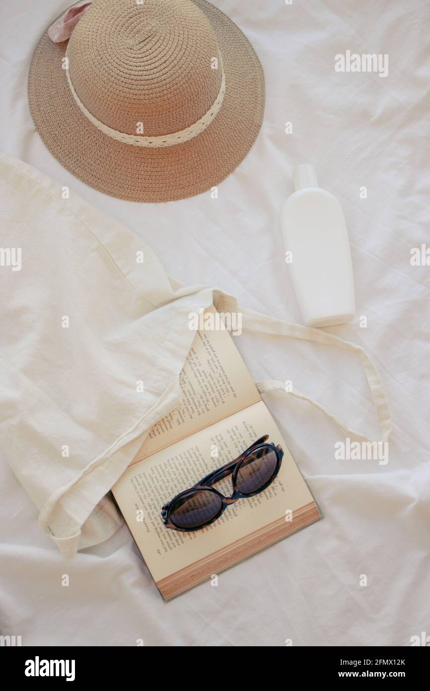 Immagine verticale di un cappello estivo, di un Sunscream, di un libro aperto e di occhiali da sole. Materiali estivi per godersi una giornata di sole. Concetto estivo 2021. Foto Stock