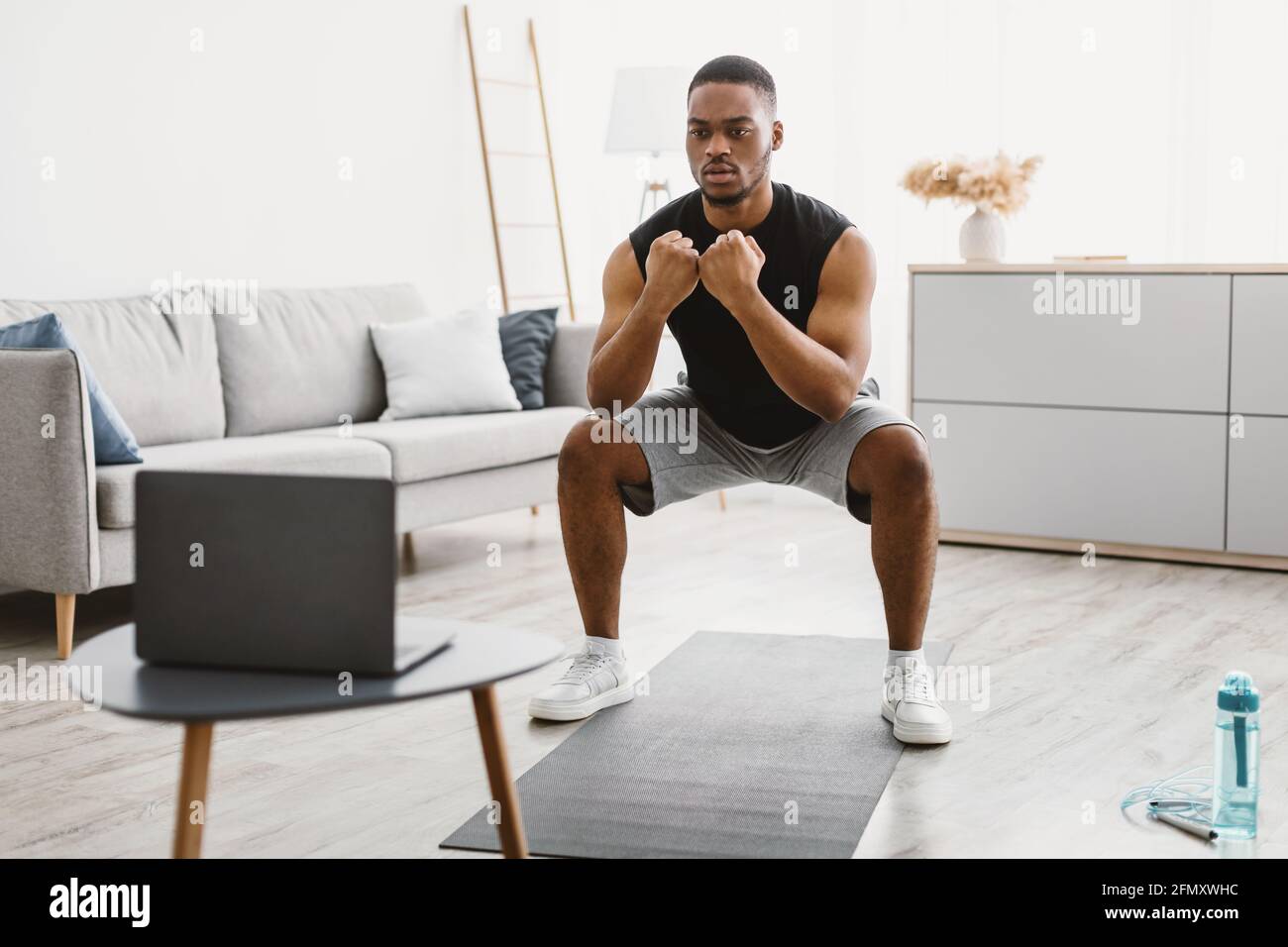 Atletico afroamericano Guy facendo squats al laptop a casa Foto Stock