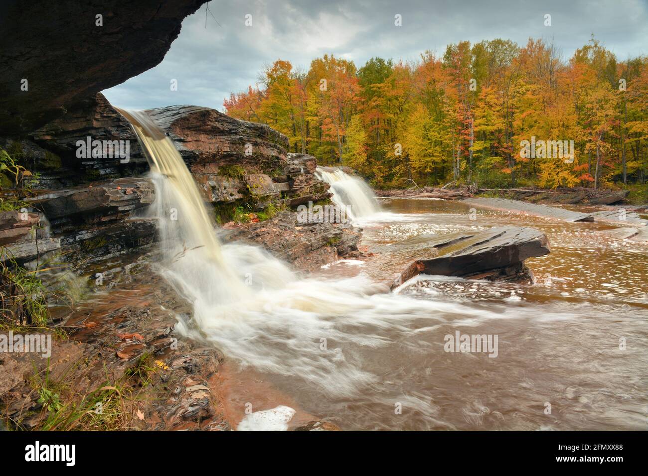 Una cascata invia sillose ruscelli a valle contro uno sfondo di boschi ricchi di colori autunnali Foto Stock