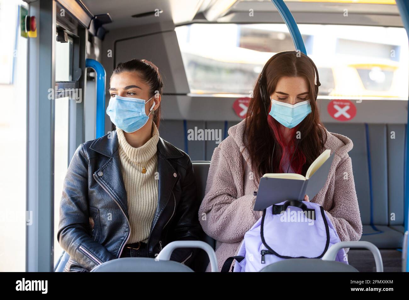 due donne che indossano una maschera protettiva seduta su un autobus uno accanto all'altro, guardando verso l'esterno la finestra lettura Foto Stock