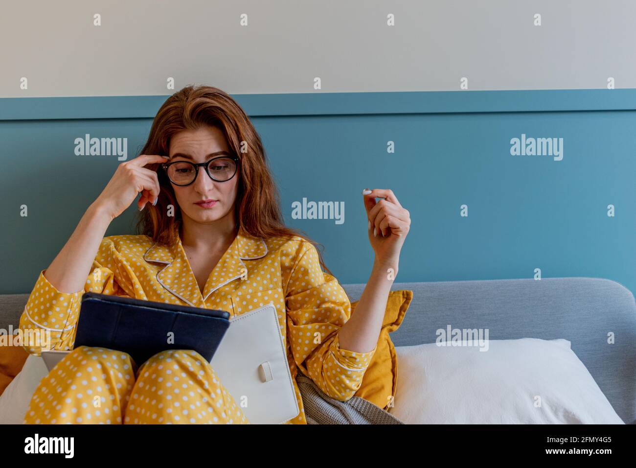 Una giovane donna con un comodo pigiama giallo lavora mentre si sdraia il letto in camera da letto. una donna d'affari mantiene un distanziamento sociale lavora a distanza. Nuovo concetto normale Foto Stock