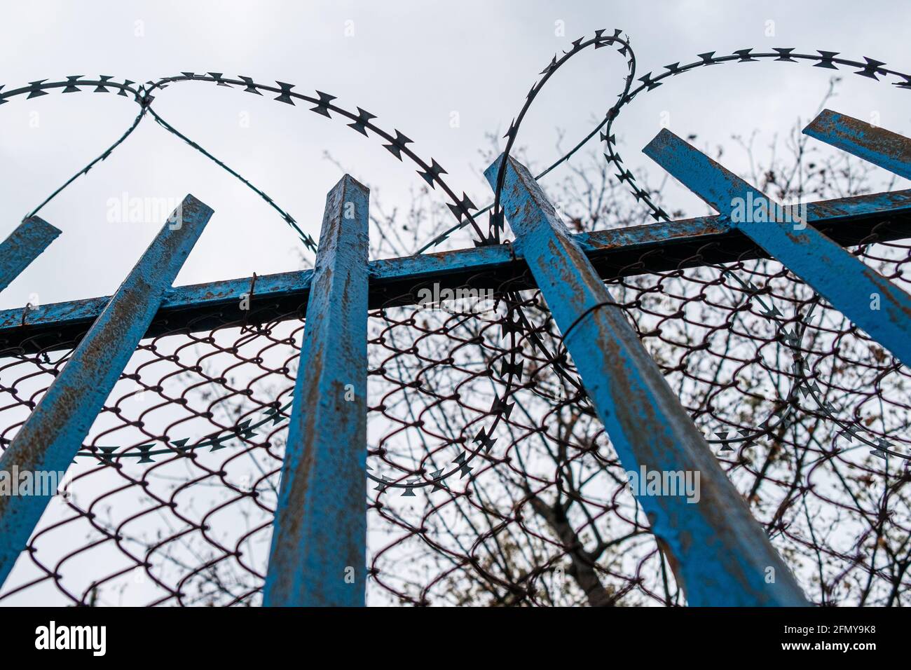 Una recinzione blu con filo spinato vicino alla prigione o all'ospedale mentale sullo sfondo del cielo grigio. Foto Stock