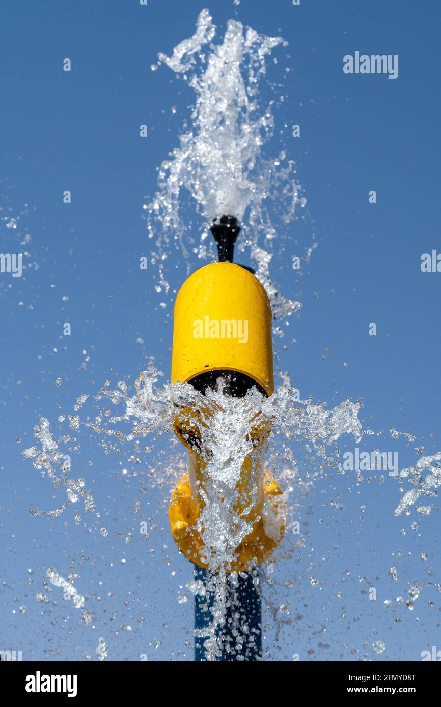 Divertimento estivo per i bambini al paraspruzzi per giocare fontana gialla spruzzando acqua al cielo blu luminoso (vista ritratto). Foto Stock