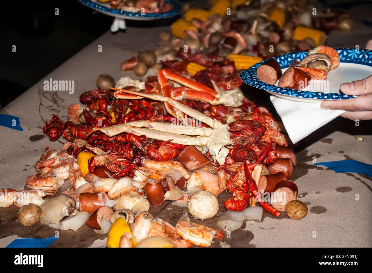 Piastra di carta con supporto a mano con pezzi di cibo selezionati da grande mucchio di frutti di mare e salsicce, patate e funghi accatastato su carta coperta tabella Foto Stock