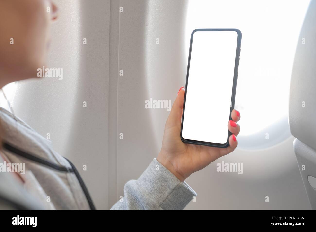 donna che tiene a mano il telefono cellulare intelligente con schermo bianco vuoto a bordo di un aereo vicino al posto della finestra. Donna che usa il telefono cellulare mentre è seduto in aereo. Foto Stock