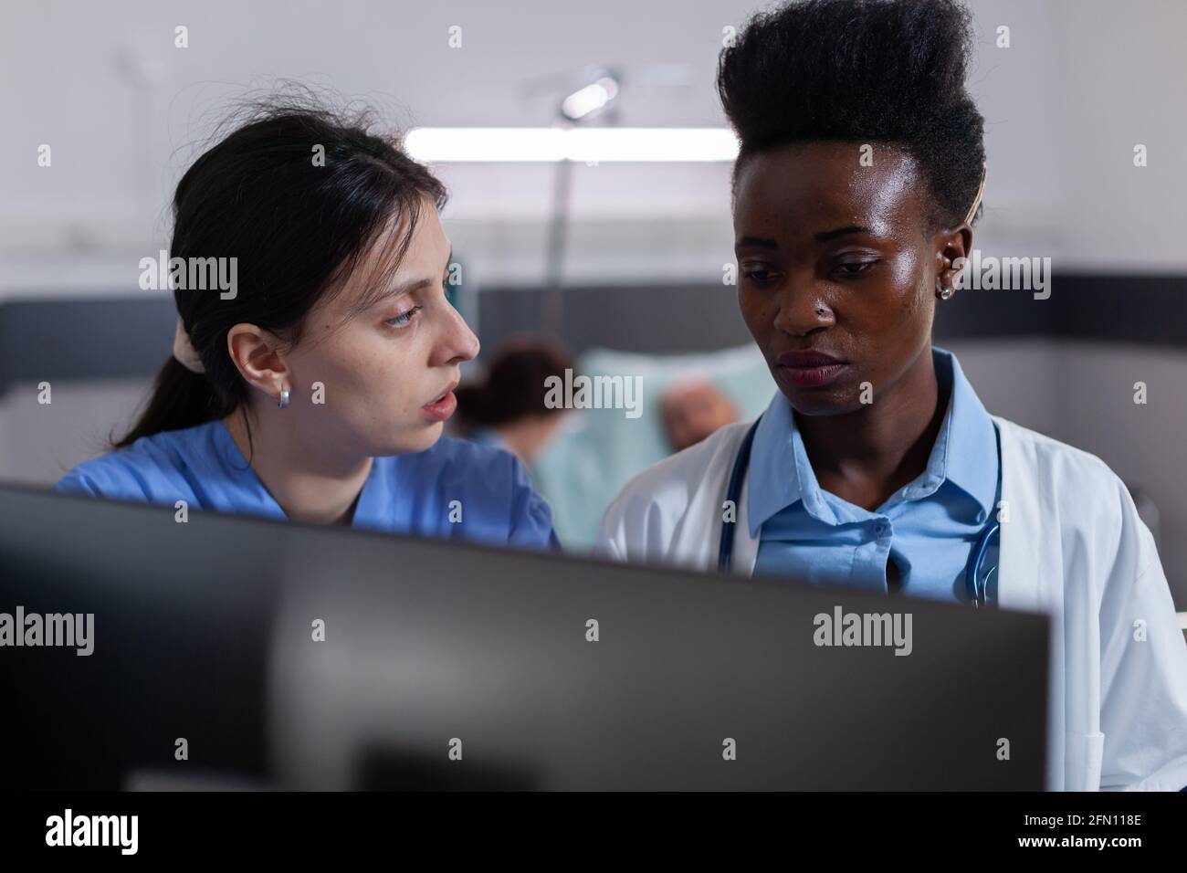 Team medico che sta esaminando il computer che analizza le cure mediche che lavorano nel reparto ospedaliero. In background medico di discutere con malato paziente, in guarigione dopo la malattia chirurgia Foto Stock