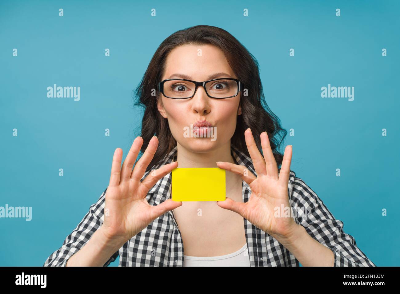 Donna sorridente carina in abito camicia e occhiali, mostrando carta di credito in mano per il concetto di società finanziaria e cashless, in posa su sfondo blu Foto Stock