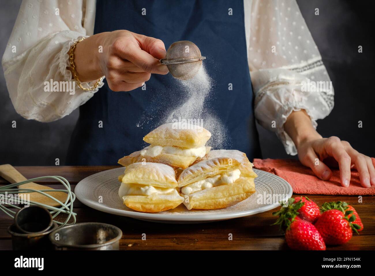 spolverare lo zucchero da un setaccio in mano alla donna, cucinando pasta sfoglia dolce con zucchero a velo alla crema Foto Stock