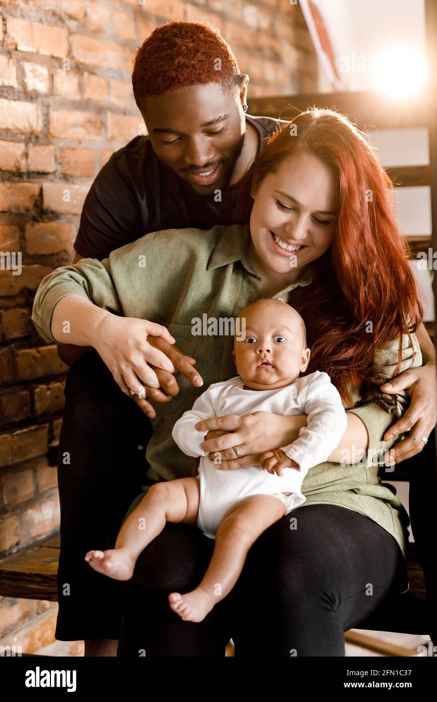 Ritratto di razza mista sorridente famiglia sedersi sulle scale. Bella madre e sorridente padre che tiene il bambino ragazzo. Genitori gioiosi che guardano con dolcezza il loro Foto Stock