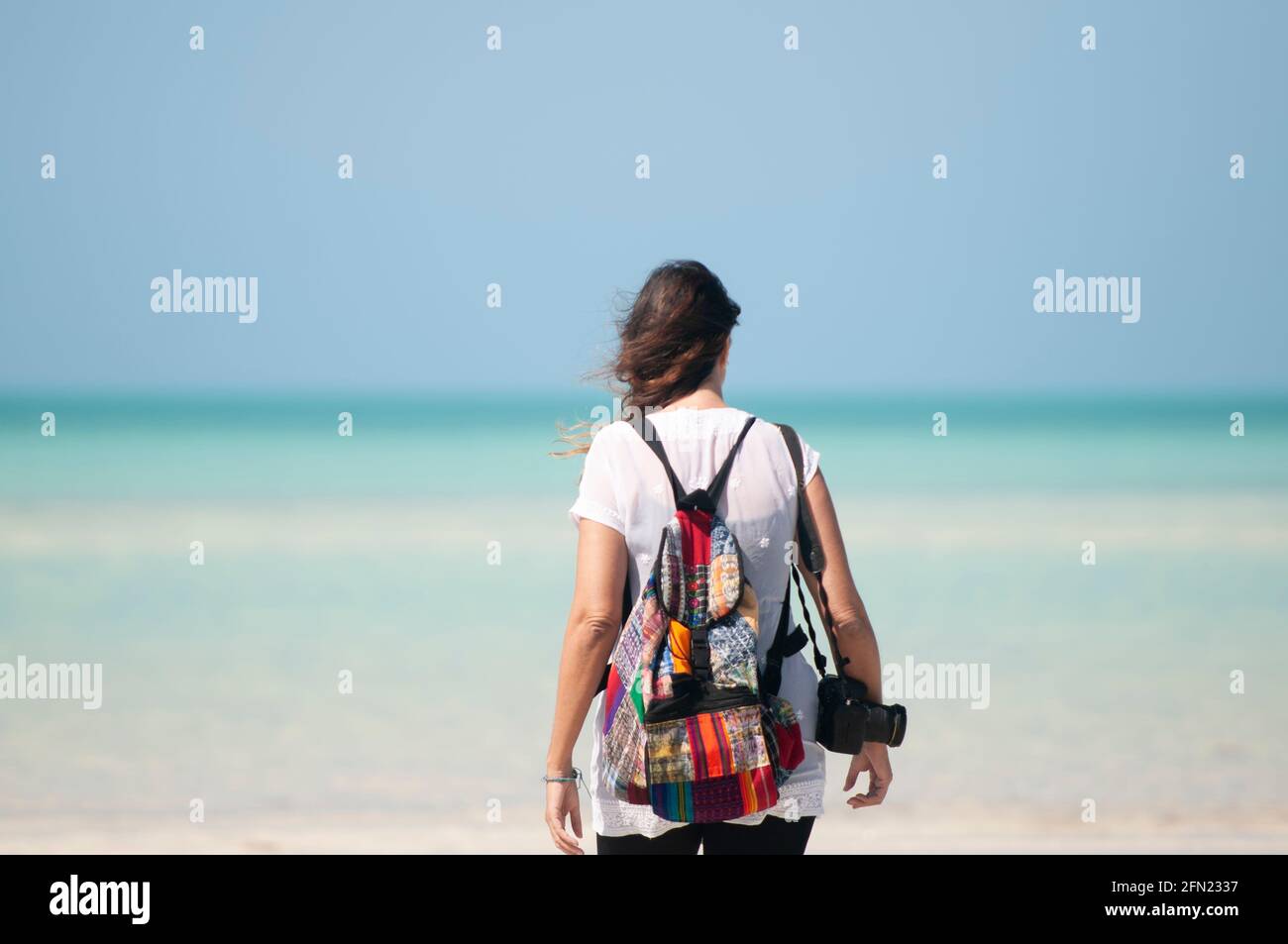 Retrovisione di una giovane donna sconosciuta che guarda verso il mare. Una fotografa con uno zaino sulle spalle e una macchina fotografica. Sullo sfondo i Caraibi messicani Foto Stock