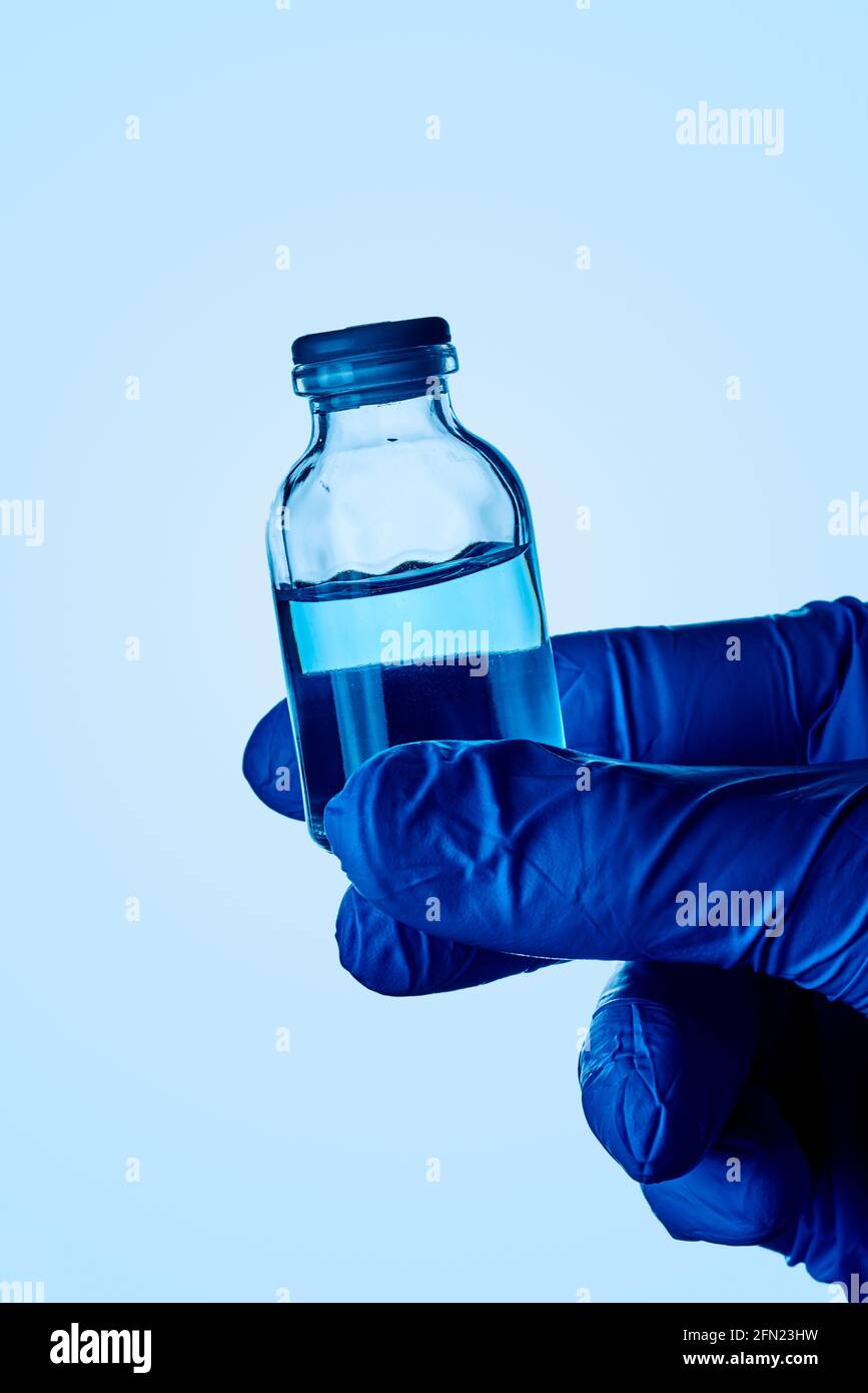 primo piano di un operatore sanitario o di laboratorio, indossando guanti chirurgici blu, con un flaconcino con un liquido blu in mano, contro uno sfondo blu chiaro Foto Stock