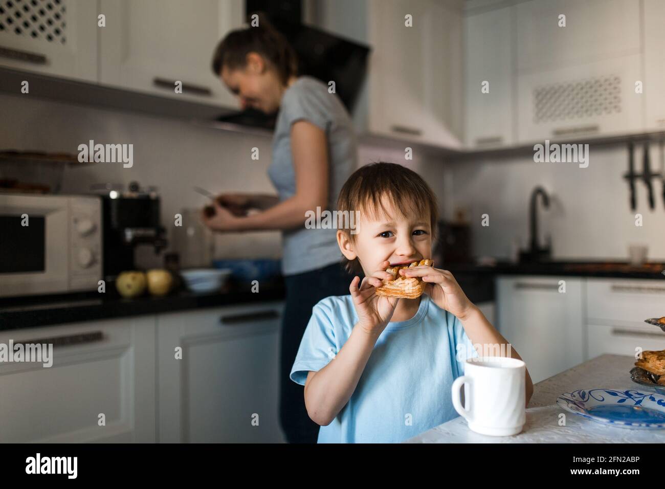 Il bambino mangia dolce torta e guarda la macchina fotografica. Background, donna prepara il cibo. Foto Stock