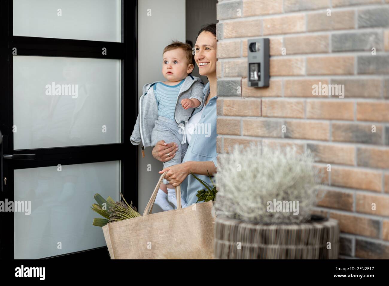 Casalinga felice con il figlio neonato che rimane di fronte alla porta della casa e portare una borsa della spesa con i prodotti dal corriere. Ordine online di cibo. Foto Stock